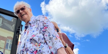 Äldre kvinna med blommig skjorta står vid en bussramp under blå himmel med vita moln.