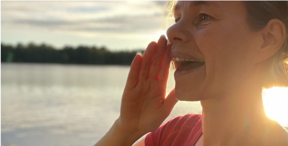 Person står vid en sjö i solnedgång och håller handen vid munnen som om de ropar.