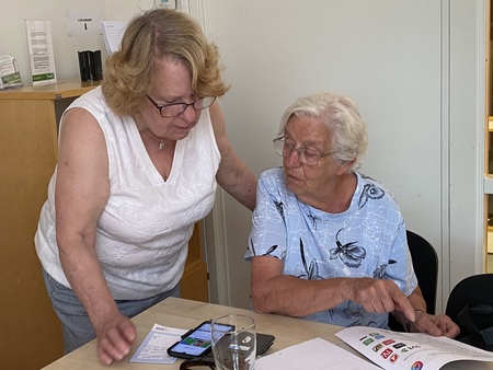 Två äldre kvinnor sitter vid ett bord och tittar på en smartphone och papper tillsammans.
