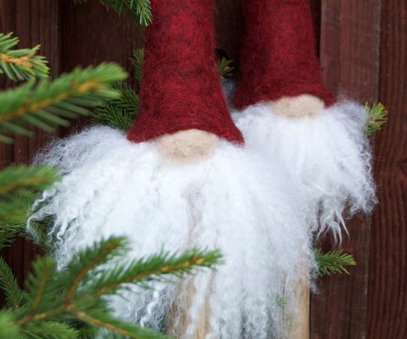 Två tomtedekorationer med vita skägg och röda luvor står bredvid en grön gran mot en trävägg.