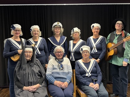 Grupp med kvinnor i sjömanskläder och en person med gitarr, poserar framför svart bakgrund.
