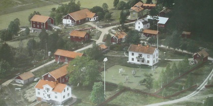 Flygfoto över en liten by med röda och vita hus, omgiven av grönska och åkrar.