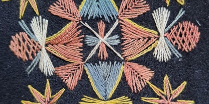 Broderi med geometriska mönster och stjärnformer i gult, rosa, blått och vitt på mörk bakgrund.