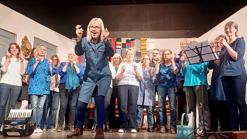En grupp kvinnor och några män står på en scen, Bodil Bendixon jeansklänning leder dem, de ser ut att sjunga eller framföra något tillsammans.