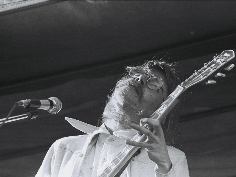 Man med långt hår och glasögon spelar elgitarr på en scen, svartvit bild.