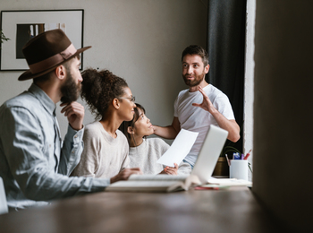 Fyra personer har ett möte vid ett bord, en man står och pratar medan de andra lyssnar.