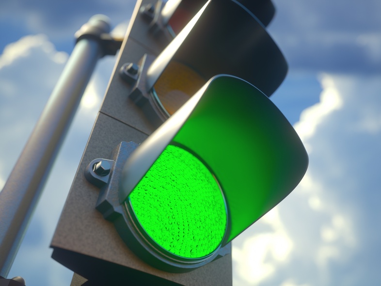 Närbild på ett trafikljus som lyser grönt mot en blå himmel med moln.