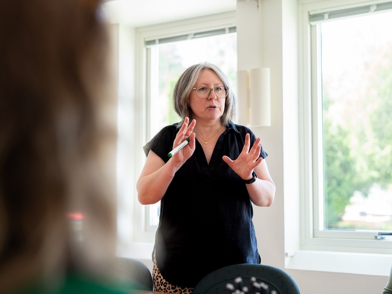 Kvinna med glasögon står och pratar framför ett fönster i ett ljust rum.