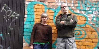 Två personer står mot en tegelvägg med färgglad graffiti, utomhus på ojämn mark.