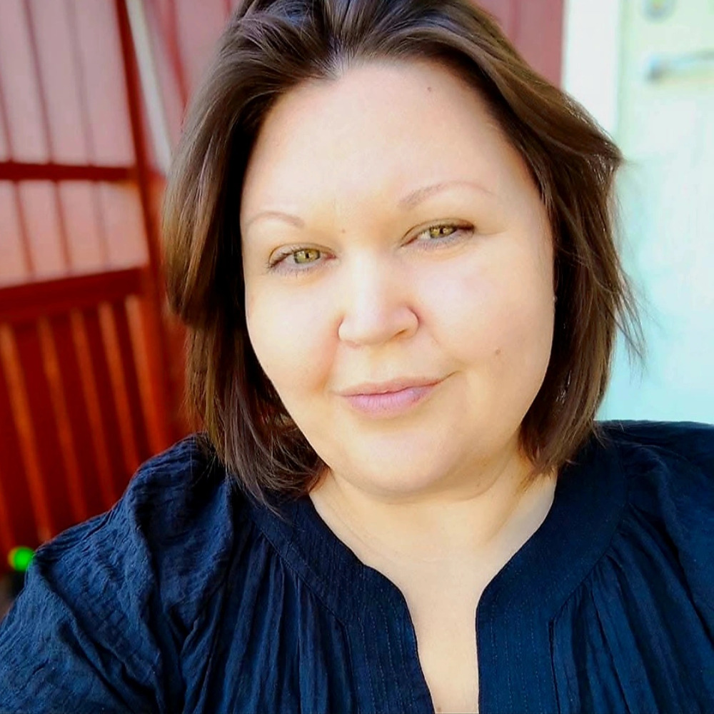 Kvinna med brunt hår och svart blus tar en selfie utomhus framför en röd vägg.