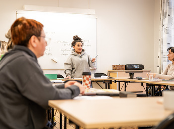 En kvinna står vid en whiteboard och undervisar en grupp vuxna i ett klassrum.
