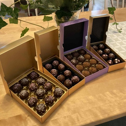 Tre öppna askar med chokladpraliner står på ett träbord bredvid en stor bukett med färgglada blommor.