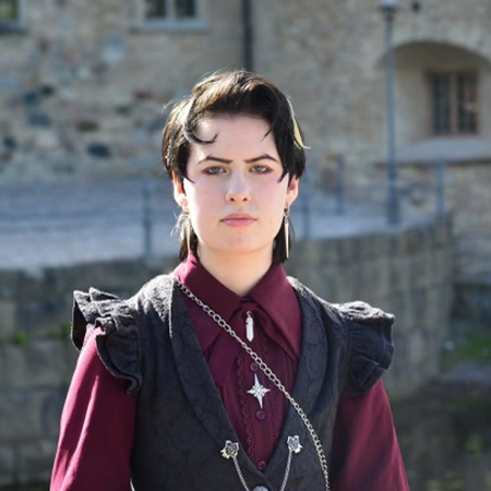 En person med kort mörkt hår och smycken bär en vinröd skjorta och svart väst, står utomhus framför en stenbyggnad.