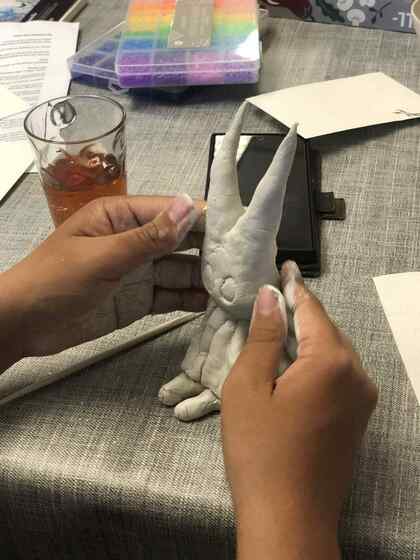 Två händer formar en kaninliknande figur av lera på ett bord med verktyg och ett glas dryck.