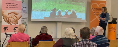 En kvinna håller en presentation för en grupp äldre personer i ett ljust konferensrum.