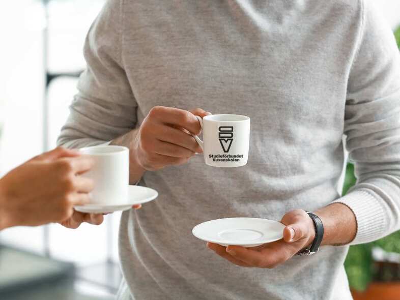 Två personer dricker kaffe ur kopp med fat. På koppen är en SV-logotyp.