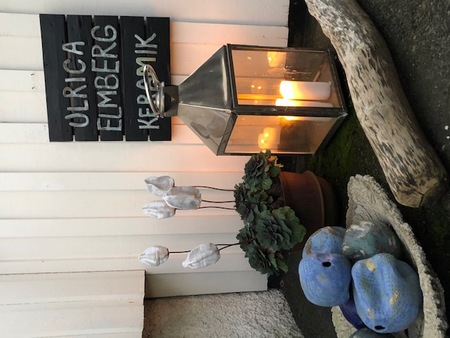 Keramikdekorationer, en lykta med tända ljus och en skylt med texten 'Ulrica Elmberg Keramik' utomhus.