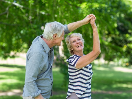 Ett äldre par dansar och skrattar tillsammans utomhus i en grönskande park.