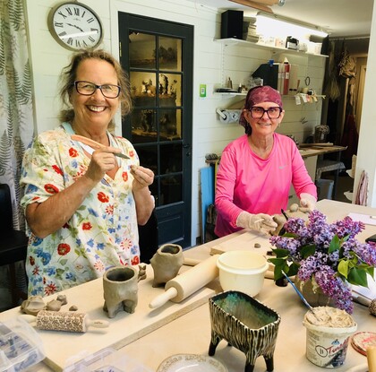 Två glada kvinnor arbetar med keramik i en ljus ateljé, omgivna av verktyg och lila blommor på bordet.