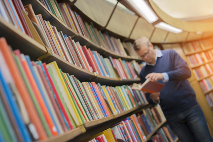 En man står i ett bibliotek och läser i en bok framför en hylla full av färgglada böcker.