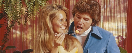 En man och en kvinna står vid ett bord fullt av frukt, mannen håller ett glas juice och båda ler.