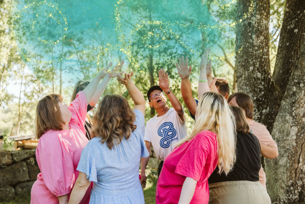 En grupp människor står i en ring utomhus och kastar färgat pulver i luften under träd.