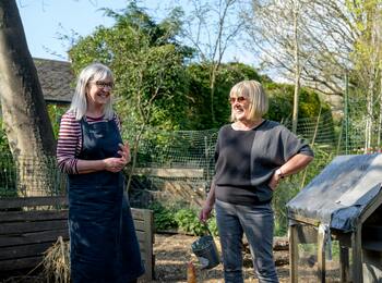 Två kvinnor samtalar i en trädgård nära ett hönshus och en kompost, med gröna träd och buskar i bakgrunden.