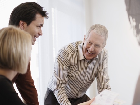 Tre personer har ett glatt samtal runt ett bord i ett ljust kontorsrum.
