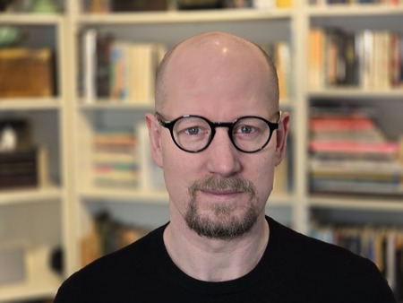 Man med glasögon och skägg står framför en bokhylla fylld med böcker.