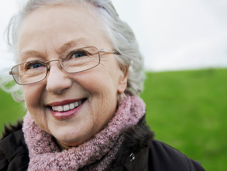 Leende äldre kvinna med glasögon och rosa halsduk utomhus på en grön äng.