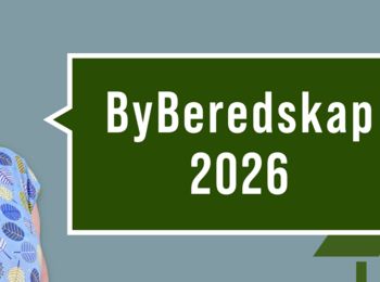 Person i blå tröja med lövmönster står bredvid en skylt med texten 'ByBeredskap 2026' mot grön bakgrund och granar.