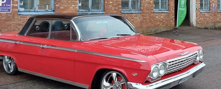 En röd klassisk bil står parkerad framför en tegelbyggnad med blå fönster.