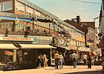Gatuliv i stadsmiljö med butiker, restaurangskyltar, flaggor och människor som promenerar en solig dag.