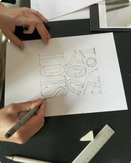 En person ritar bokstäver och mönster på ett vitt papper med en svart penna vid ett skrivbord.