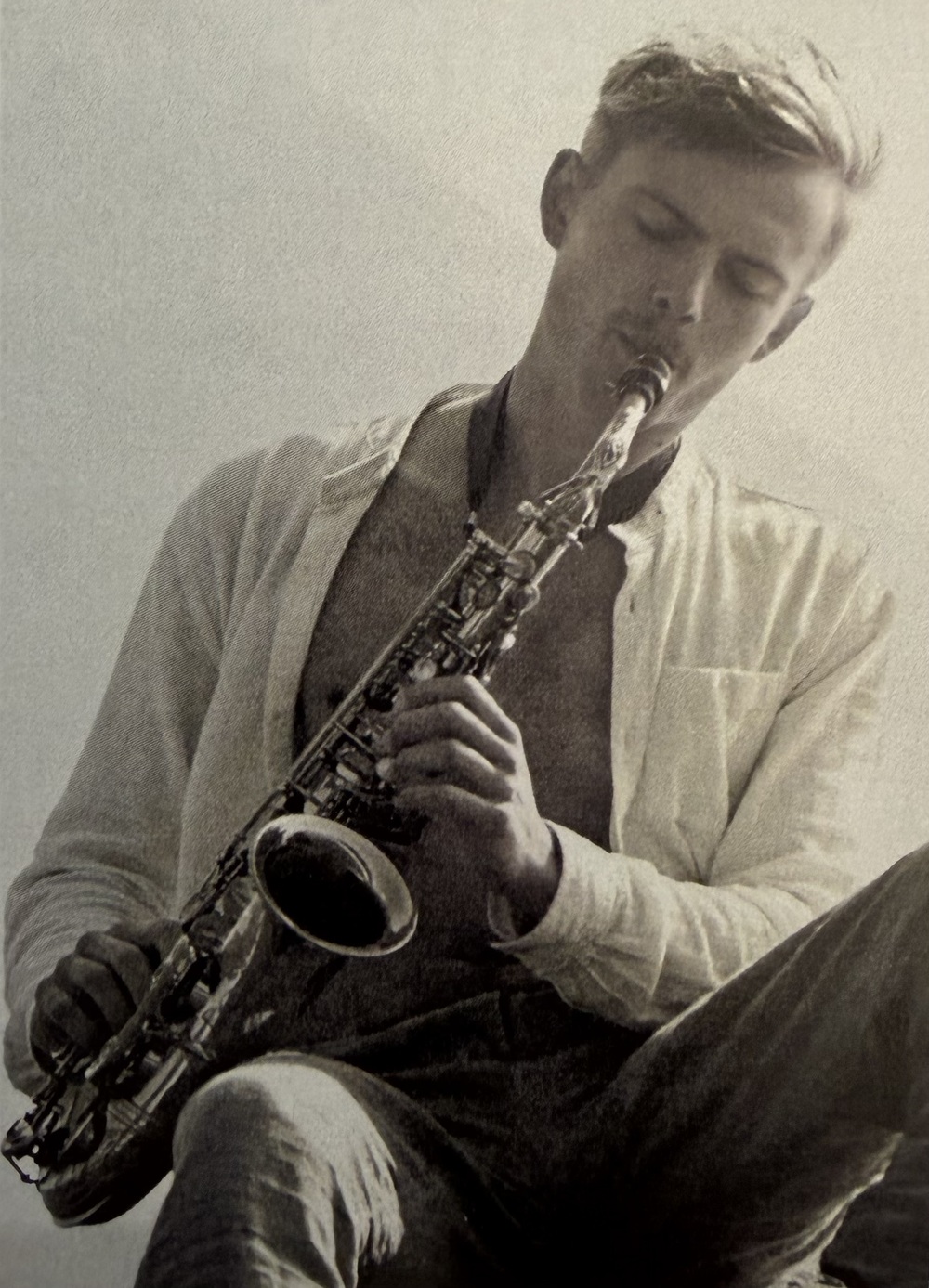Person i ljus skjorta spelar saxofon, ansiktet är suddat, svartvit bild.