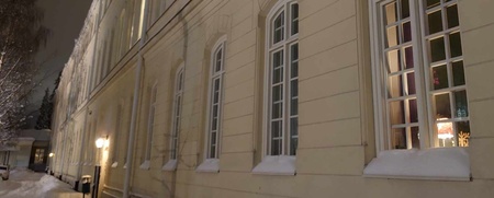 Gul byggnad med höga fönster, snö på marken och kvällsbelysning utomhus.