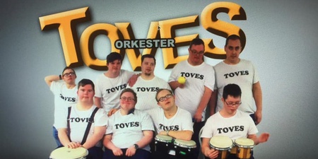 Gruppfoto av åtta personer i vita TOVES-tröjor med trummor framför en stor skylt med texten TOVES ORKESTER.