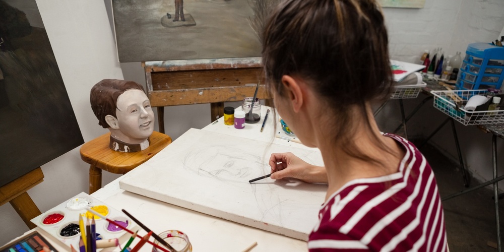 En kvinna tecknar ett porträtt vid ett konstbord med färgpennor och penslar i en ateljé.