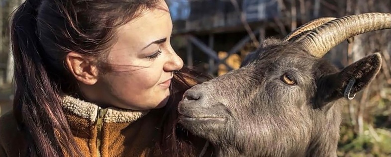 En kvinna i brun fleecejacka tittar leende på en get som står nära henne utomhus.