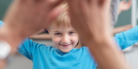 Leende barn i blå tröja sträcker ut armarna i klassrum, händer syns i förgrunden.