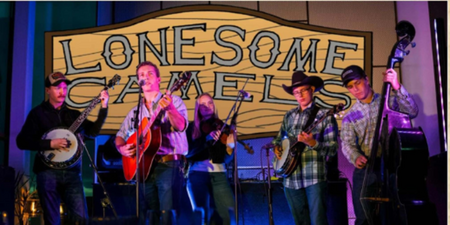 Ett bluegrassband spelar live på scen med instrument framför en skylt där det står 'Lonesome Camels'.