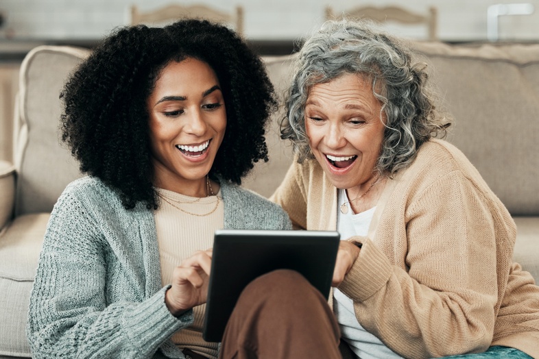 Två kvinnor sitter på golvet framför en soffa och tittar glatt på en surfplatta tillsammans.