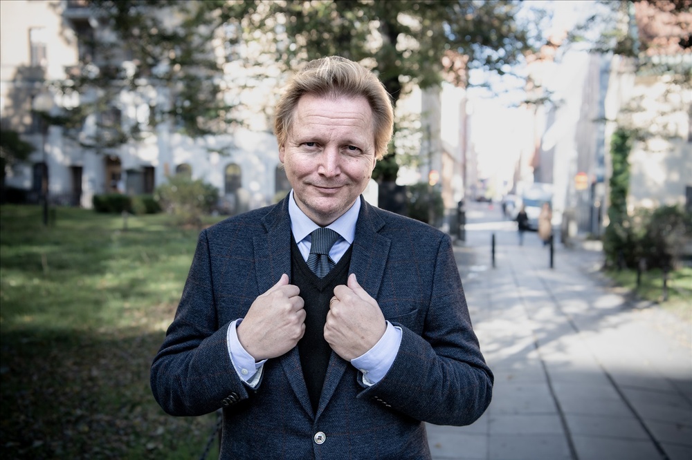 Man i kostym står utomhus på en trottoar med händerna på kavajen och ler mot kameran.