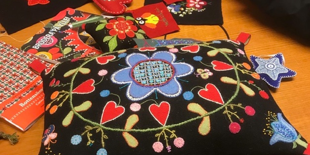 Två personer arbetar med färgglad broderi på svart tyg vid ett bord, med kuddar och tygbitar.