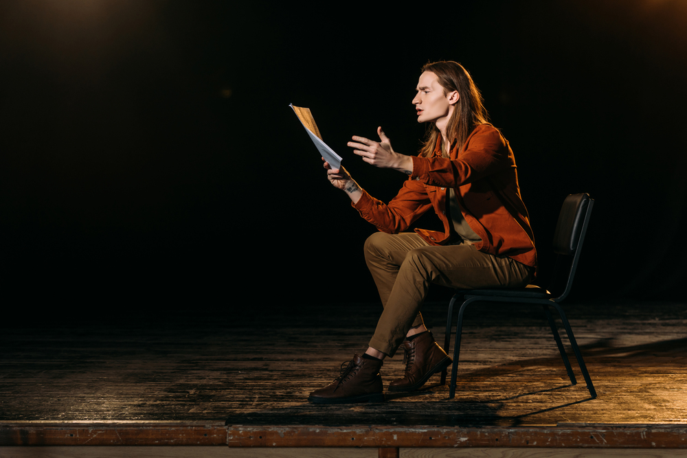 En person sitter på en stol på en scen och gestikulerar med ett manus i handen under en teaterövning.