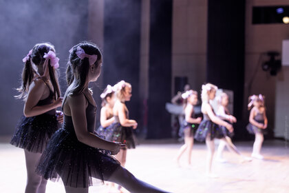 Flera unga flickor i svarta balettklänningar dansar på en scen med ljus bakgrund.