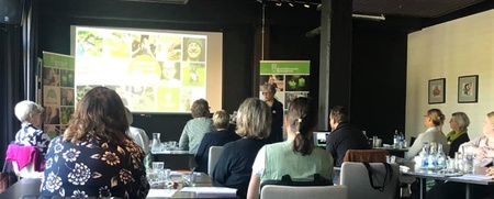 En grupp människor sitter vid bord och lyssnar på en presentation i ett konferensrum.