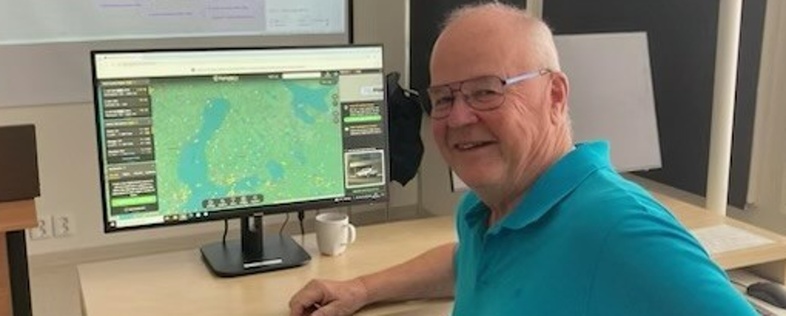 Äldre man i blå tröja sitter vid skrivbord med dator och karta på skärmen, ler mot kameran.