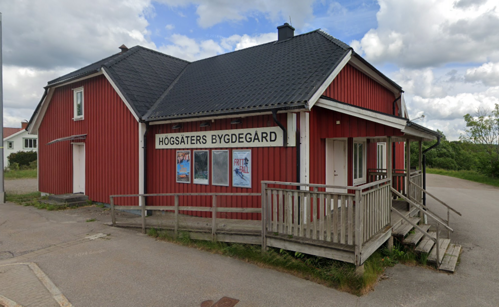 Röd träbyggnad med skylt 'Högsäters Bygdegård' och ramp vid entrén, landsbygdsmiljö.