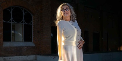 Kvinna i ljus kjol och tröja står på en perrong med tegelvägg och stora fönster i bakgrunden.